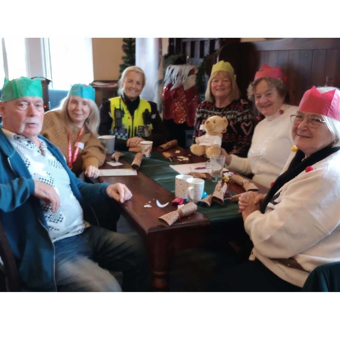 A group of six elderly people wearing colorful paper hats sits around a table, enjoying a festive gathering. - Home Instead
