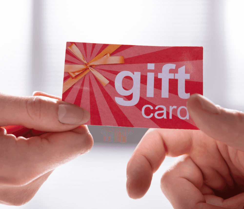 Close-up of two hands exchanging a red gift card with a yellow bow and the words "gift card" printed on it. - Home Instead