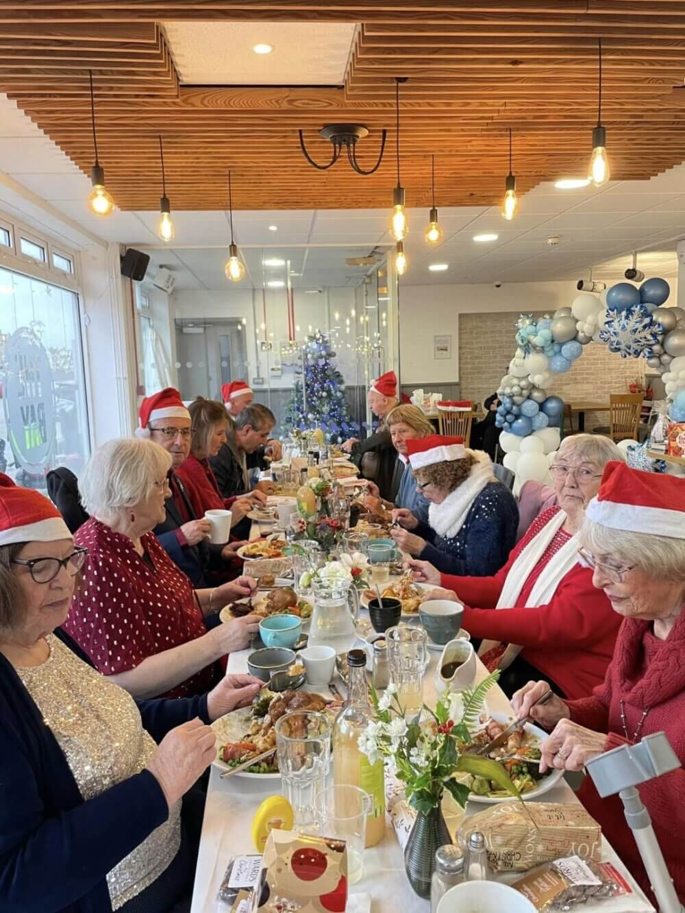 A group of people wearing Santa hats are seated at a long table, enjoying a meal in a festive, decorated room. - Home Instead