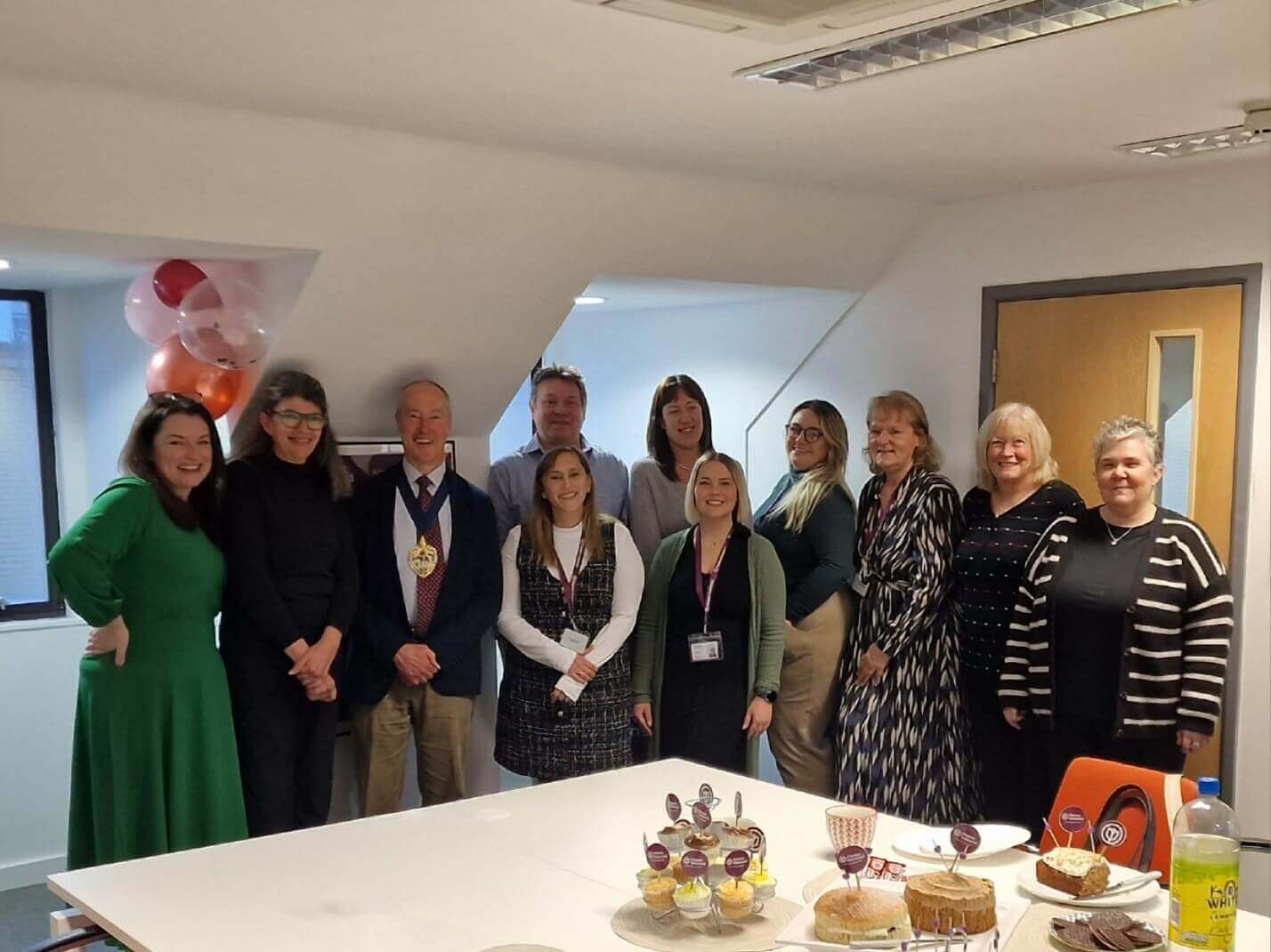 A group of people standing together in an office, with a table of cupcakes and cakes in the foreground. - Home Instead