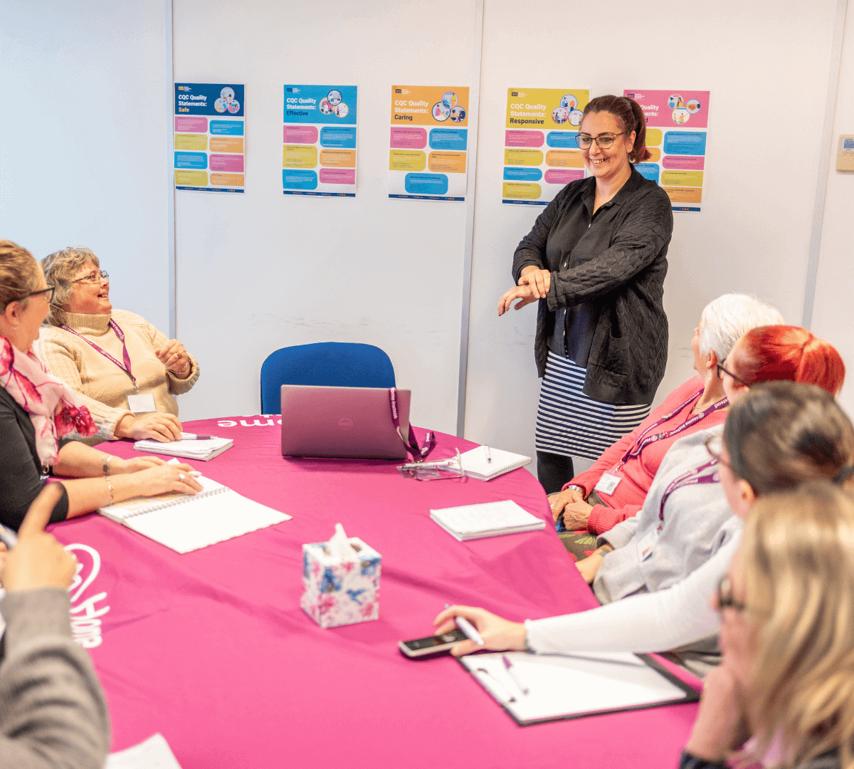 A woman is standing and gesturing to a group of seated people during a meeting in a brightly lit room. - Home Instead