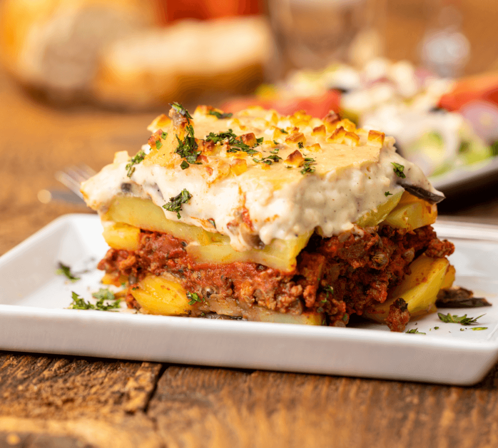 A slice of moussaka on a white plate, garnished with parsley, with a blurred salad in the background. - Home Instead