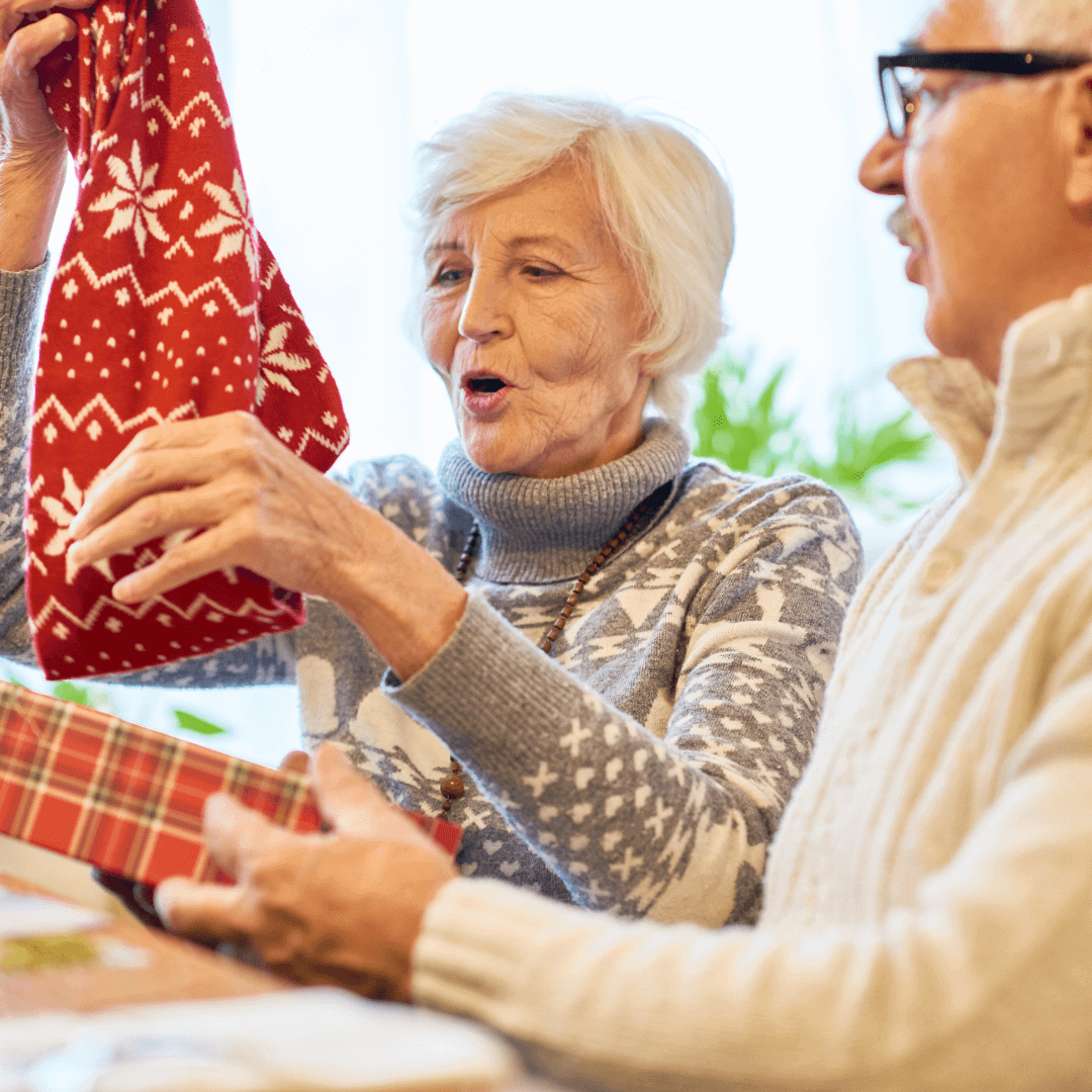 An elderly woman excitedly pulls a gift from a Christmas stocking while an elderly man watches and smiles. - Home Instead