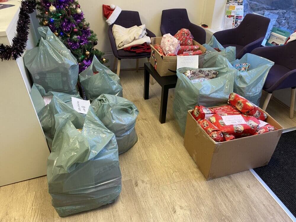 A room with chairs, plastic bags, and a box, all filled with wrapped Christmas gifts next to a decorated tree. - Home Instead