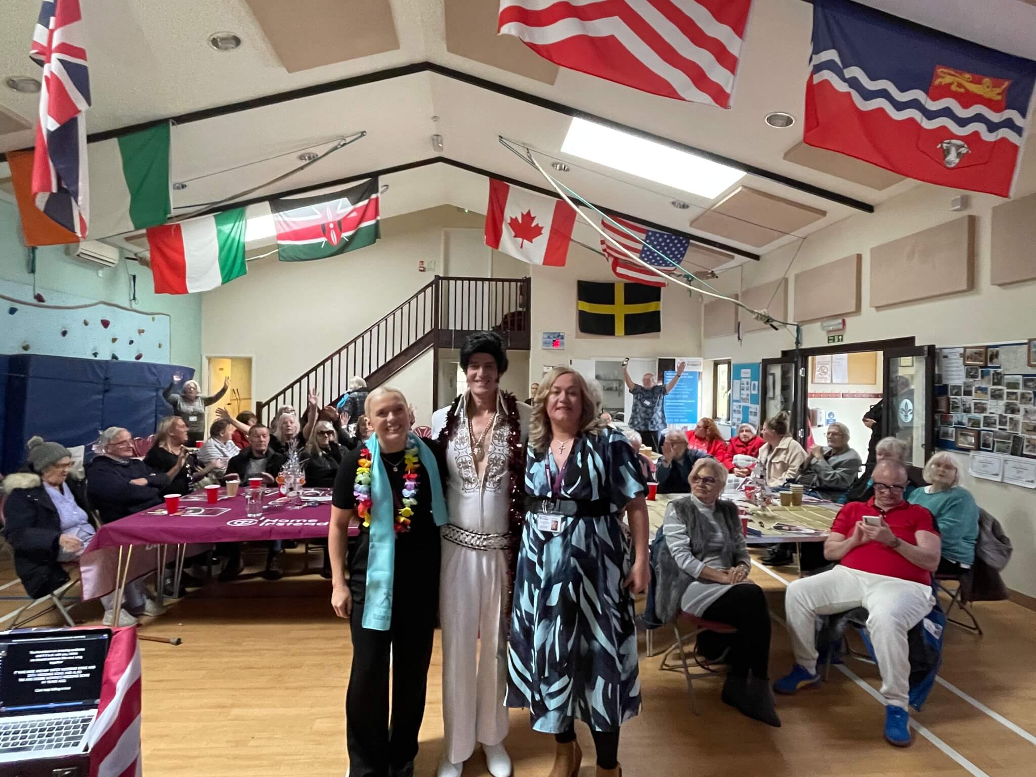 A group of people in a community hall decorated with international flags, centered around a performer in a white outfit. - Home Instead