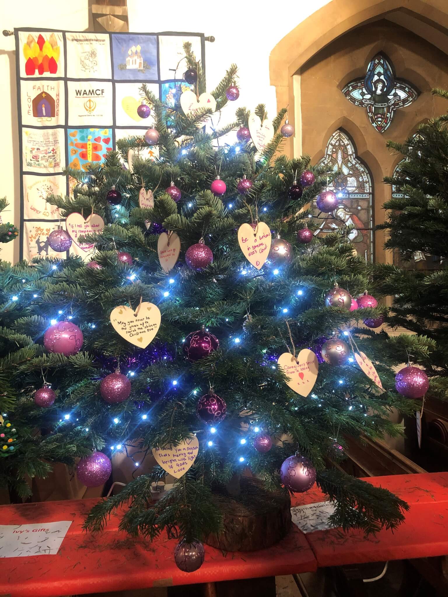 Christmas tree decorated with purple ornaments, heart-shaped tags, and blue lights, situated in a festive indoor setting. - Home Instead