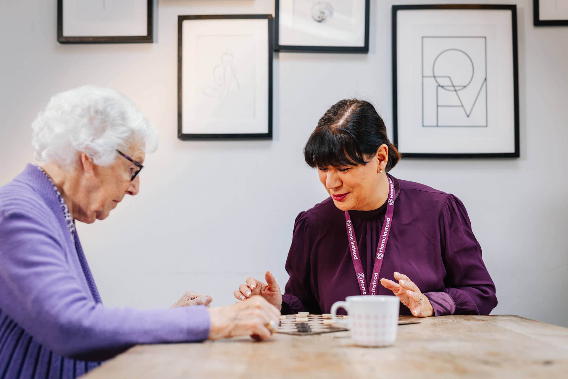 Elderly woman playing a board game with a caregiver wearing a purple top, sitting at a wooden table with framed photos behind. - Home Instead
