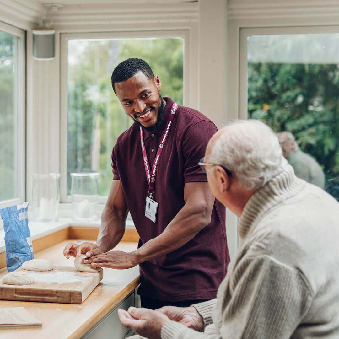 A smiling caregiver kneads dough at a kitchen counter while an elderly man watches and smiles. - Home Instead