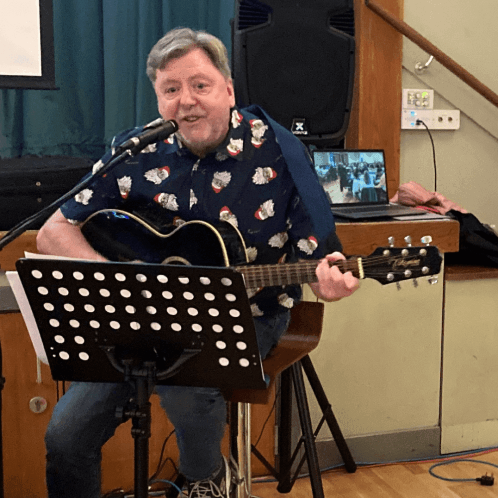 A man in a festive shirt plays an acoustic guitar while singing into a microphone, with a music stand in front of him. - Home Instead