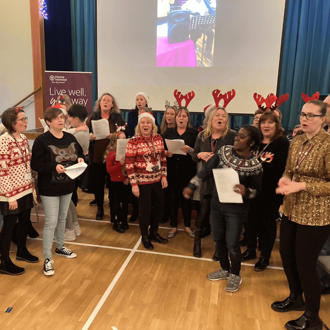 A group of people wearing festive outfits sing together on a wooden floor with a projector screen in the background. - Home Instead