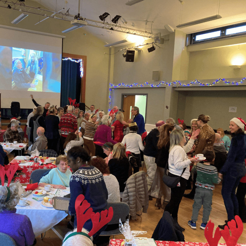 People at a festive indoor gathering, many wearing holiday attire and reindeer antlers, enjoying food and socializing. - Home Instead