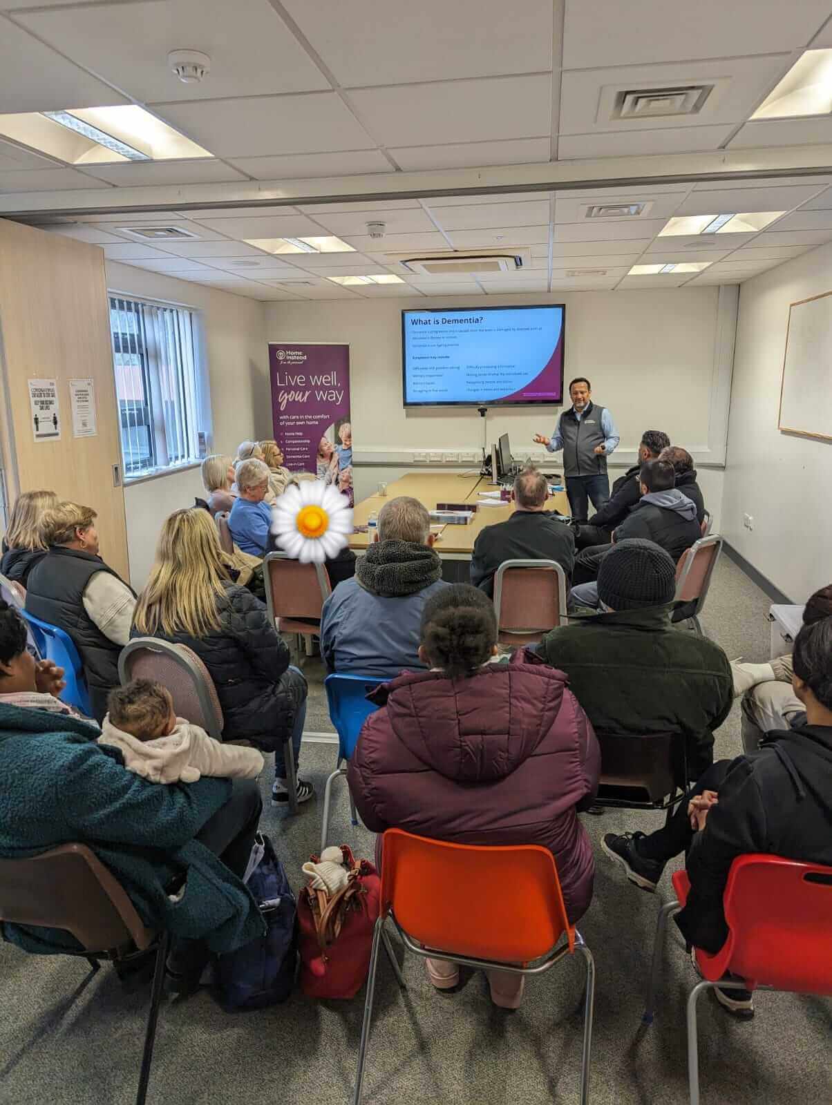 A man speaks to a seated audience in a brightly lit room with a screen displaying a presentation. - Home Instead