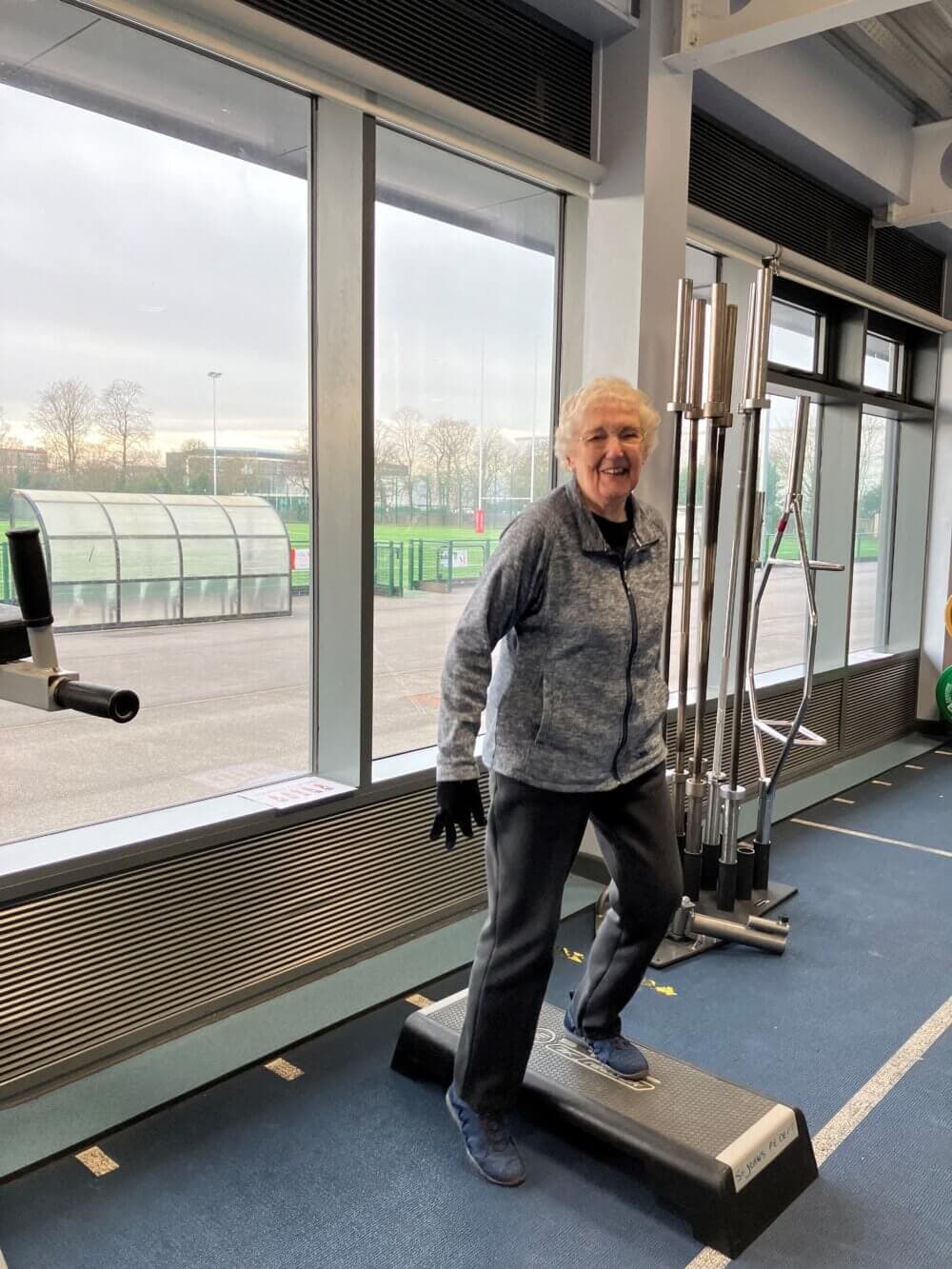 Smiling older woman exercising on a step platform in a gym with large windows and weightlifting equipment. - Home Instead
