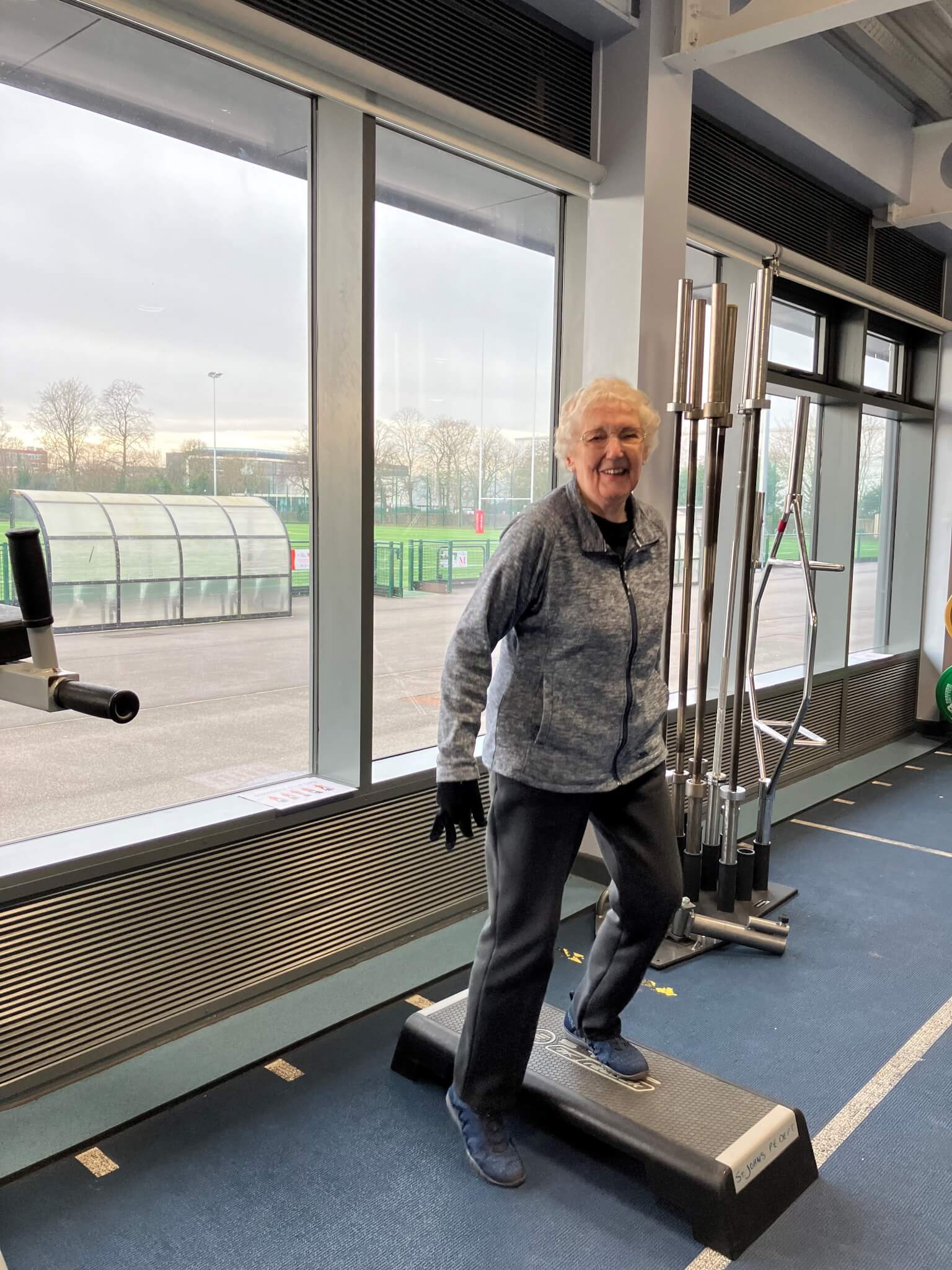 Smiling older woman exercising on a step platform in a gym with large windows and weightlifting equipment. - Home Instead