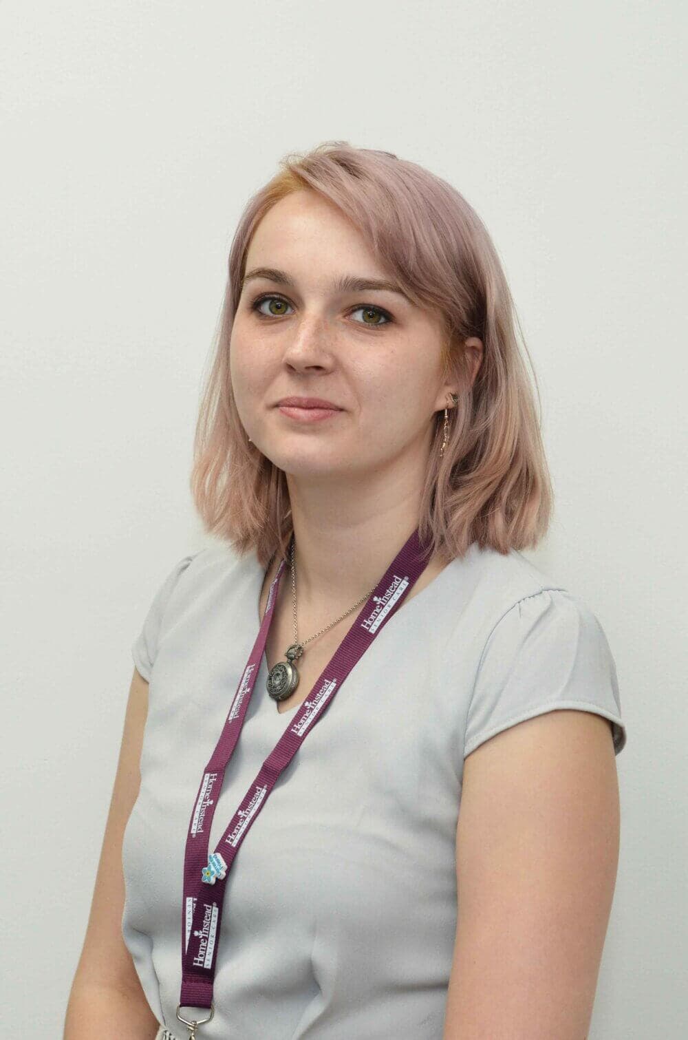 Woman with shoulder-length pink hair, wearing a light grey top and a purple lanyard, against a white background. - Home Instead