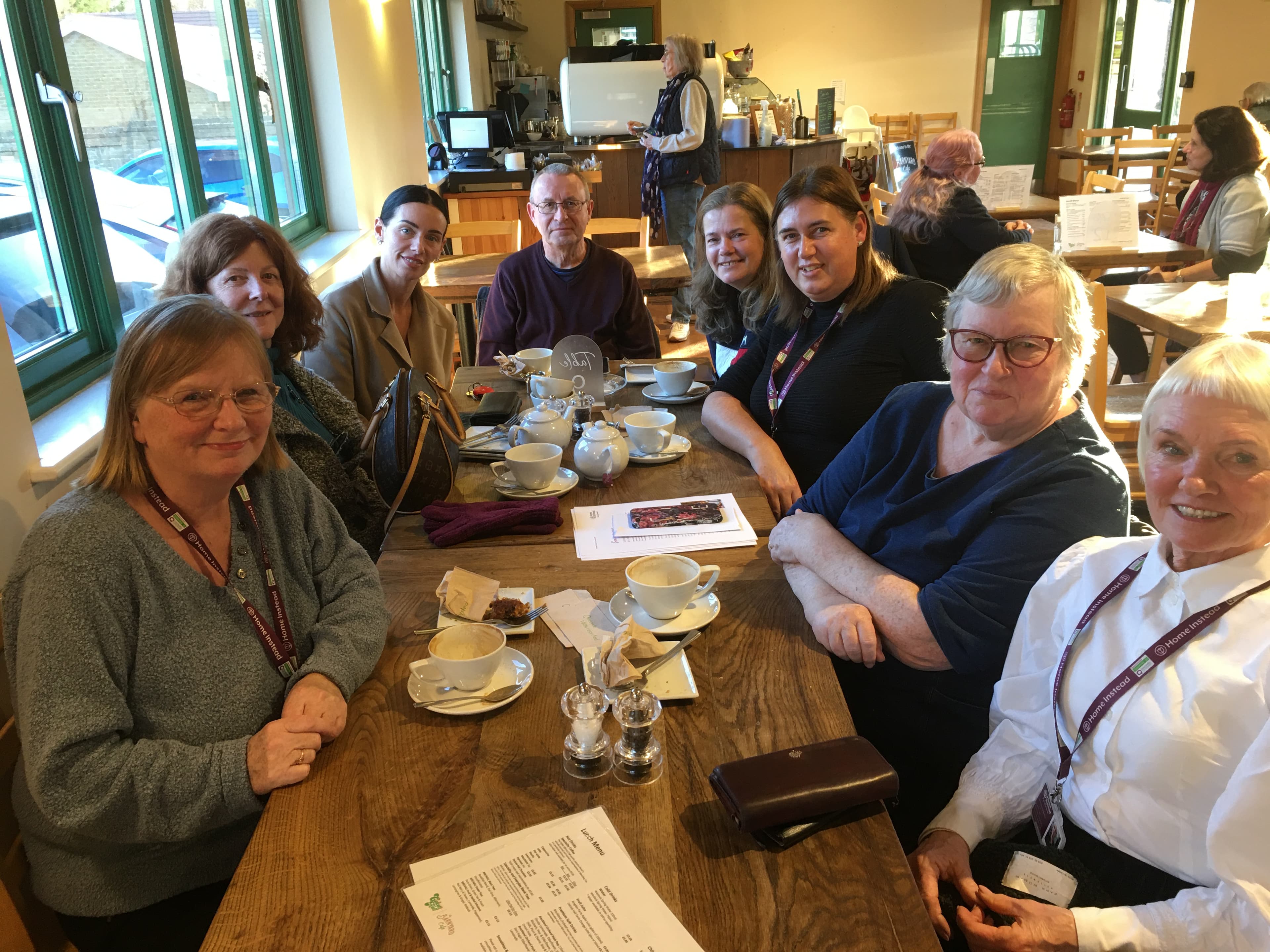 A group of people sit around a table in a cafe, smiling, with coffee cups and a menu on the table. - Home Instead