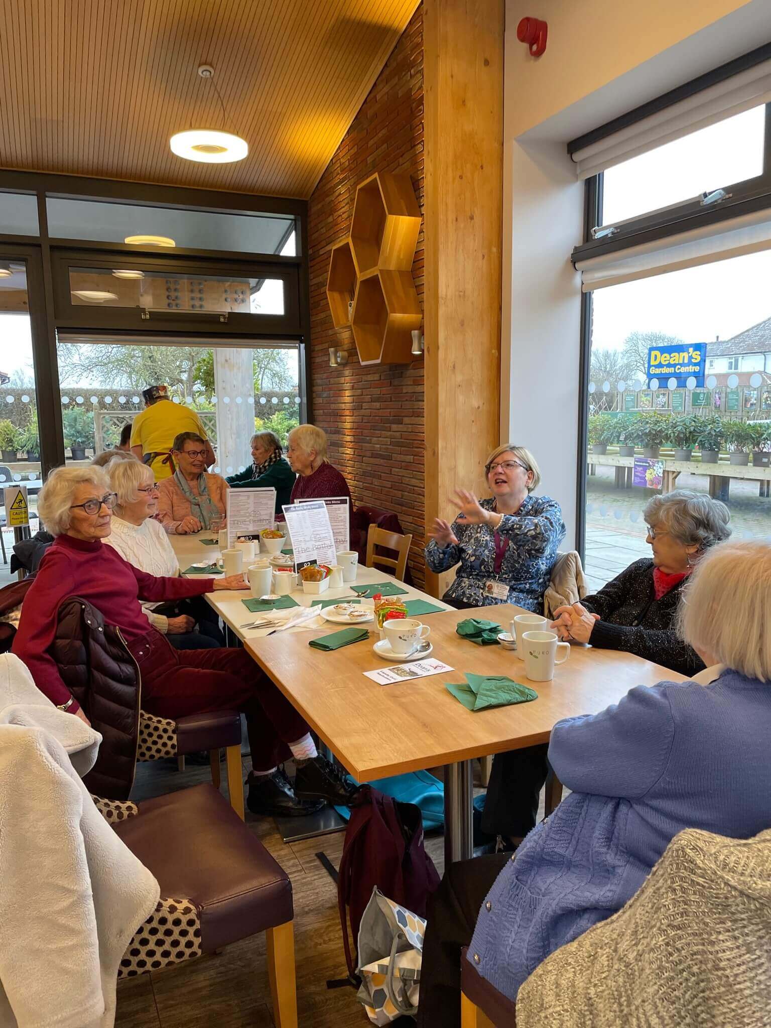 A group of elderly people sit and chat around a table in a cozy café, enjoying a meal together. - Home Instead