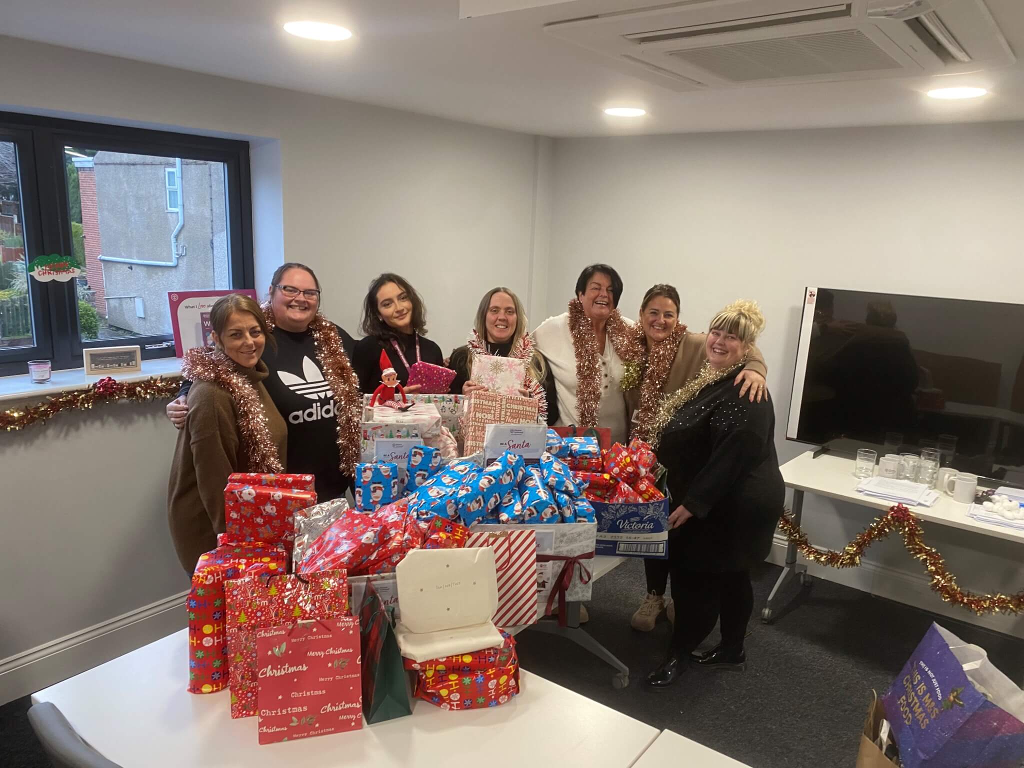 A group of seven people in a festive office setting, holding wrapped gifts and wearing tinsel scarves. - Home Instead