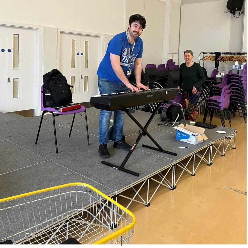 A man stands on a stage playing a keyboard while smiling, with a woman seated nearby and chairs in the background. - Home Instead