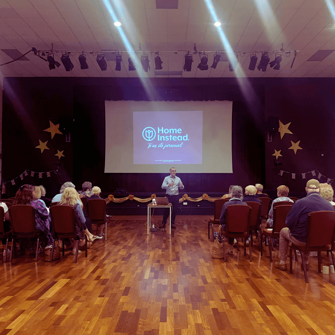 A person speaking to an audience in a hall with a "Home Instead" presentation on a screen behind them. - Home Instead