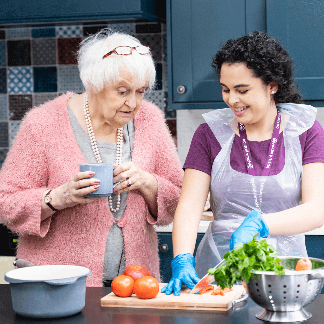 An elderly woman with a mug watches a caregiver chopping vegetables in a kitchen. Both are smiling. - Home Instead