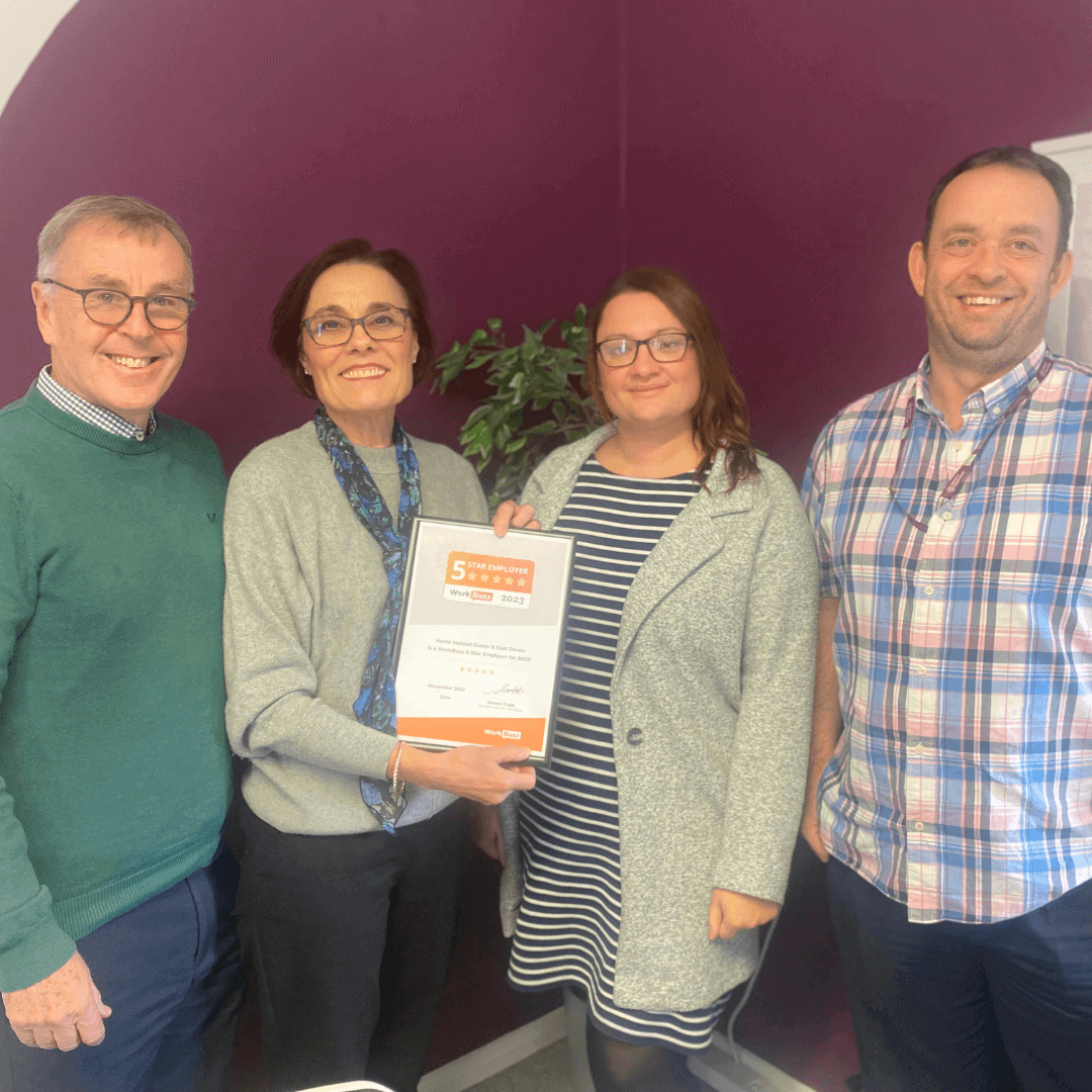 Four smiling people pose together, holding a framed certificate against a purple wall with a plant in the background. - Home Instead