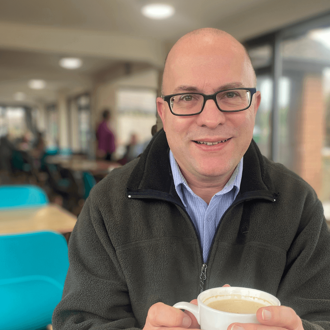 A man with glasses holds a coffee cup in a brightly lit café with turquoise chairs in the background. - Home Instead