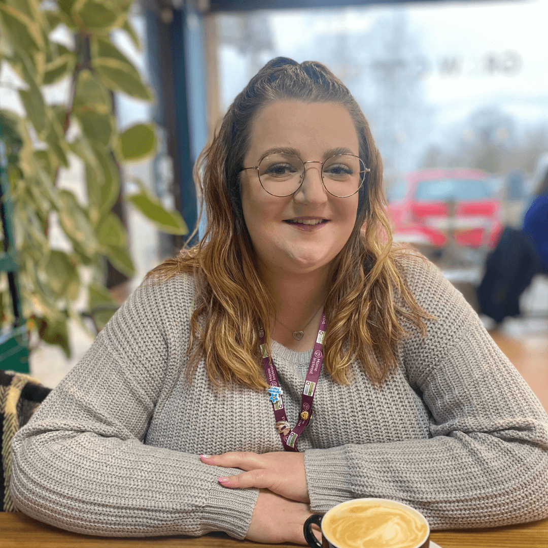 A person with glasses and wavy hair smiles while sitting at a table with a latte in a cozy indoor setting. - Home Instead