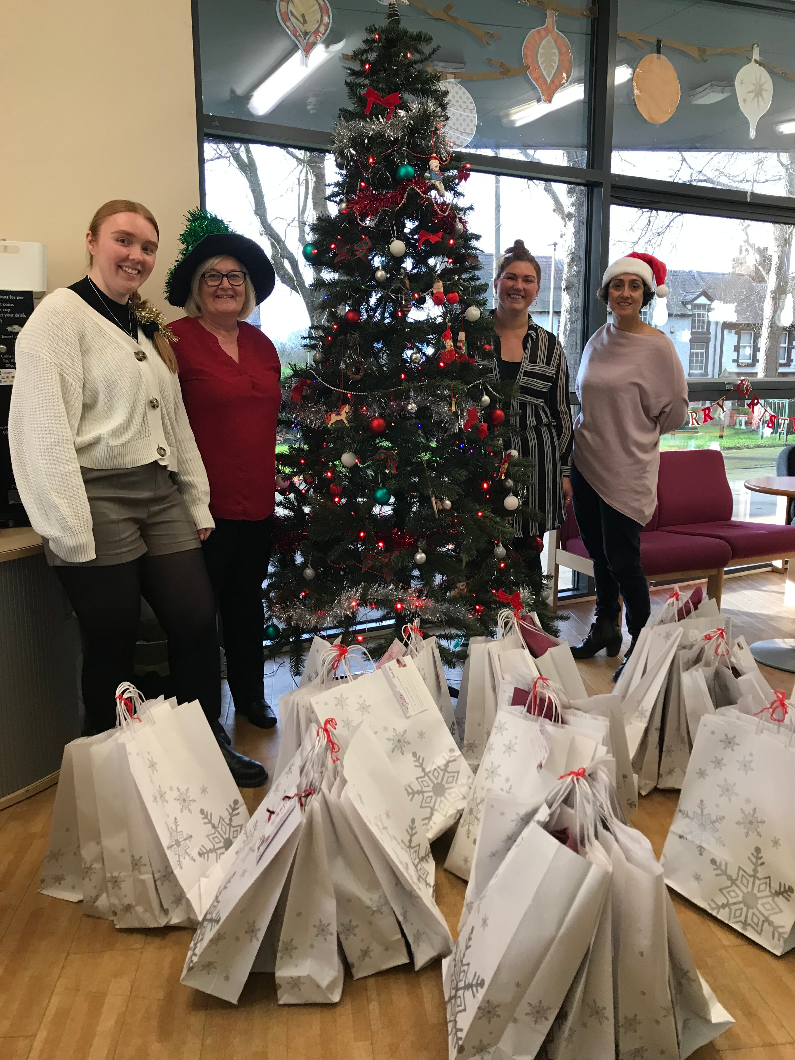 Four people stand by a decorated Christmas tree with numerous gift bags on the floor around them. - Home Instead