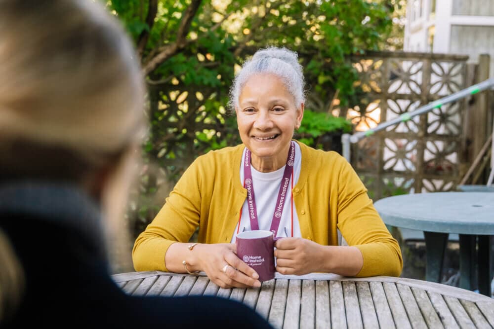 A smiling woman with gray hair, wearing a yellow cardigan and lanyard, holds a mug while sitting at a wooden table outdoors. - Home Instead