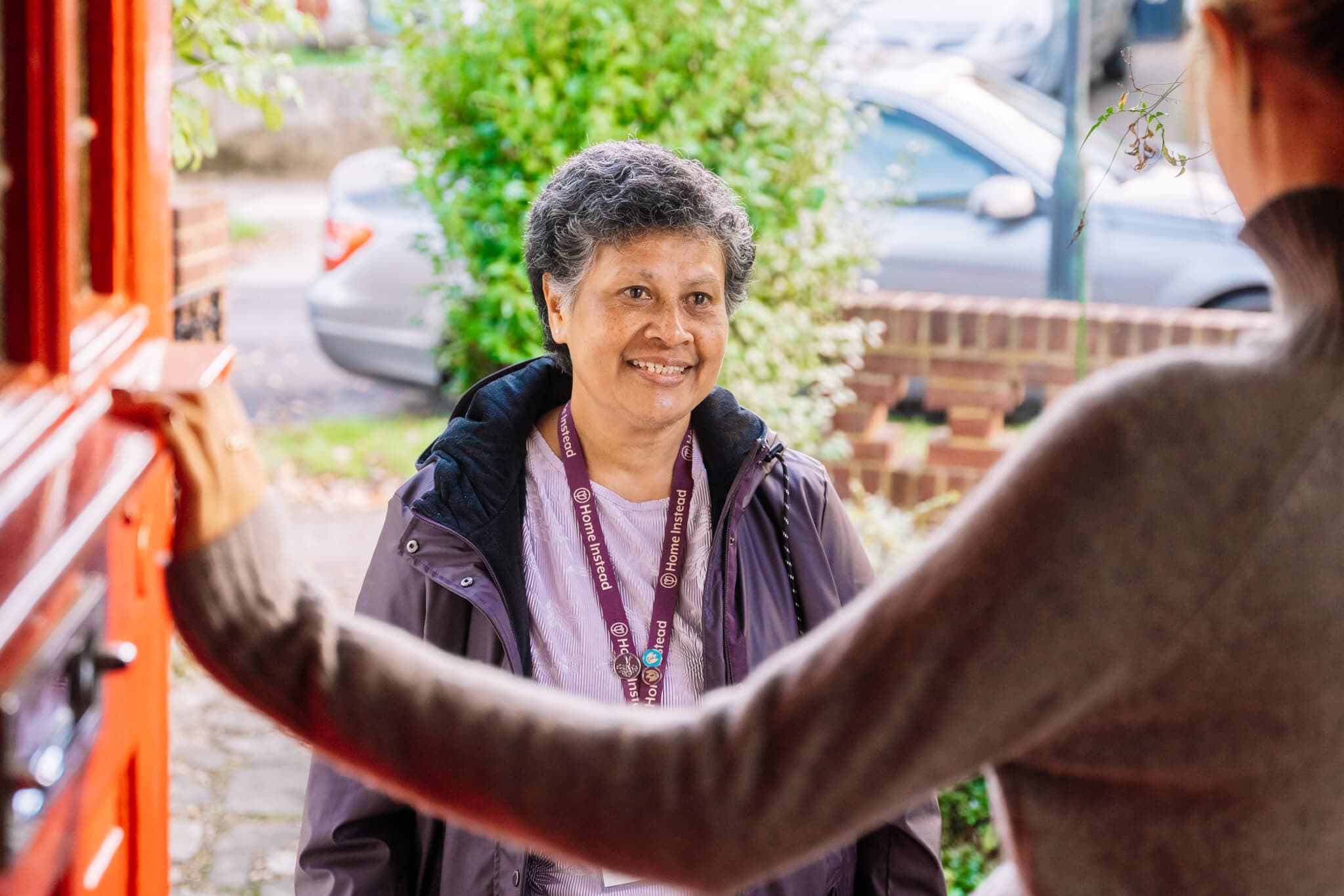 A woman smiles while visiting a person at their front door, with a car and greenery in the background. - Home Instead