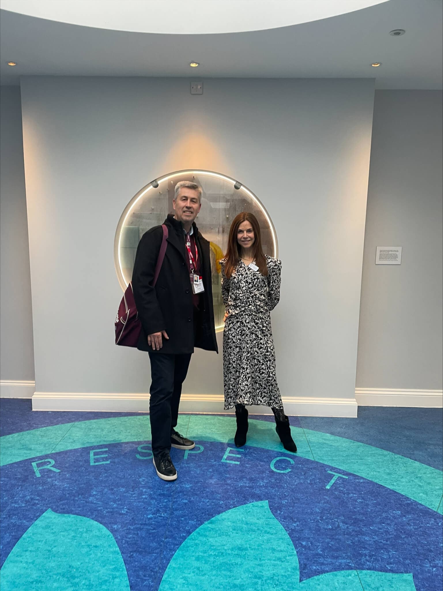 Two people stand smiling in front of a circular window. The word "RESPECT" is visible on the floor beneath them. - Home Instead