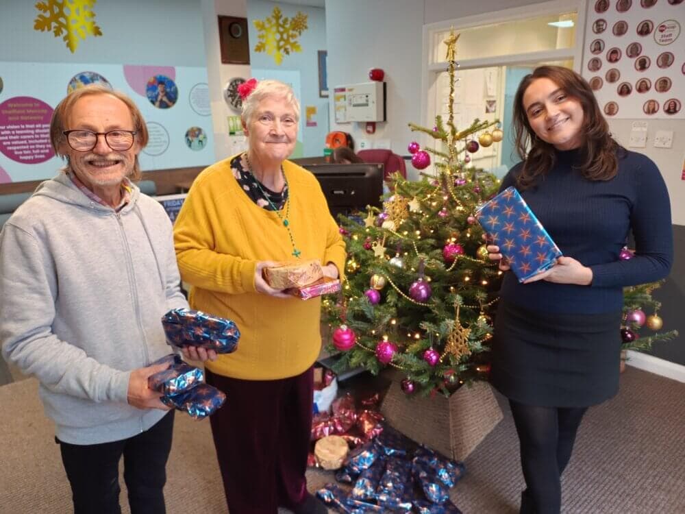 Three people stand by a decorated Christmas tree holding wrapped presents, smiling and looking at the camera. - Home Instead