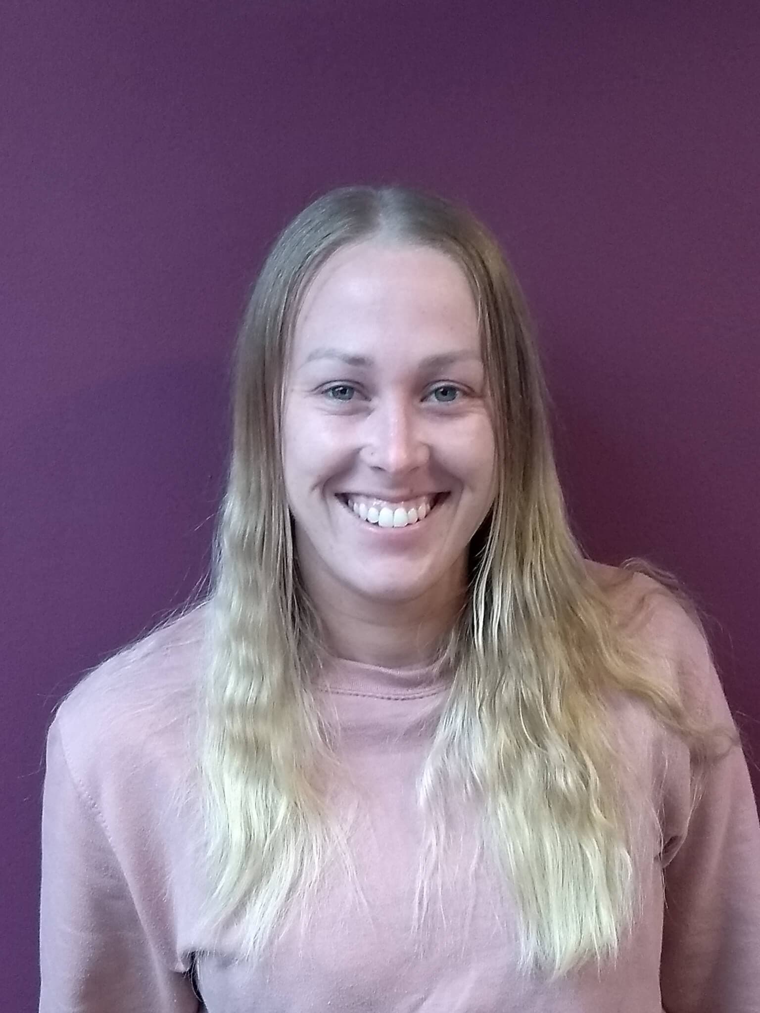 A woman with long blonde hair smiles in front of a purple background. She is wearing a light pink top. - Home Instead