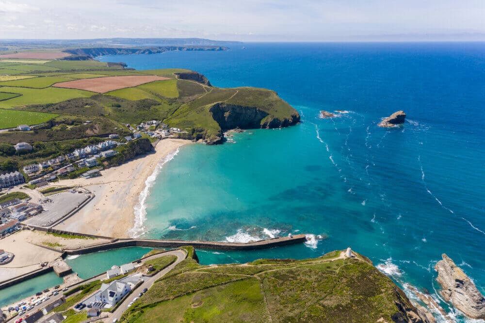 Aerial view of a coastal village with sandy beach, turquoise waters, rocky cliffs, and surrounding green fields. - Home Instead
