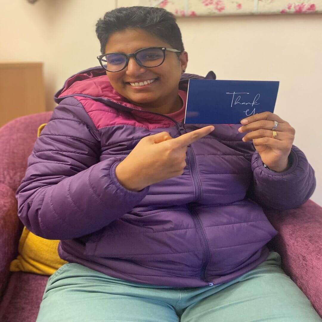 A person smiling and pointing to a blue "Thank You" card. They are wearing glasses and a purple jacket. - Home Instead