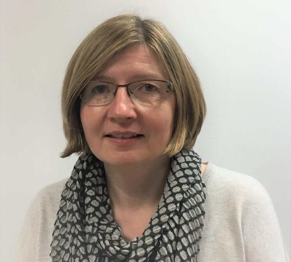 A woman with glasses and short blond hair wearing a white shirt and patterned scarf, standing in front of a white background. - Home Instead