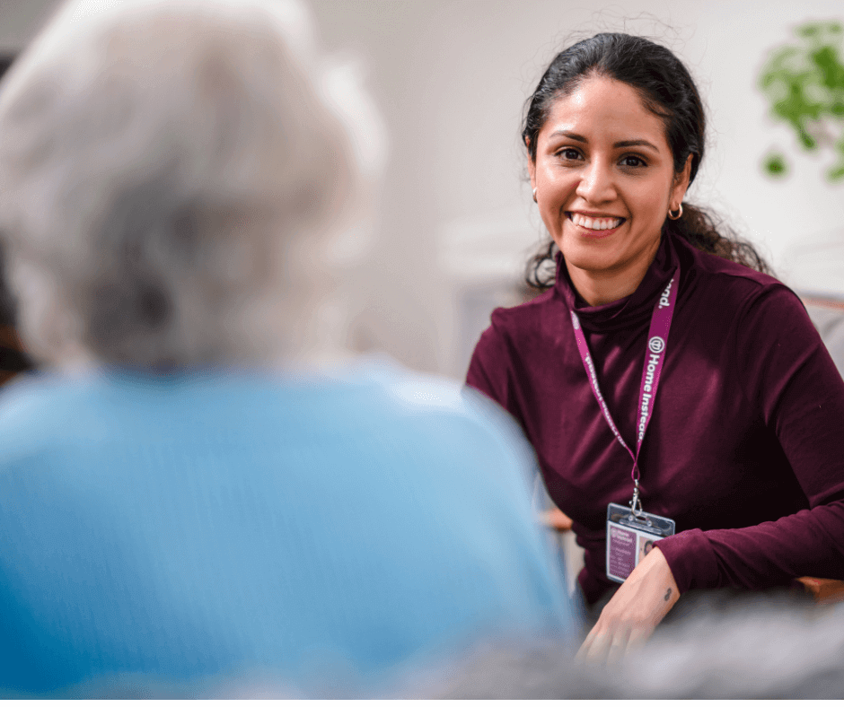 A woman in a maroon top smiles while talking to an elderly person with white hair, wearing a lanyard and name badge. - Home Instead