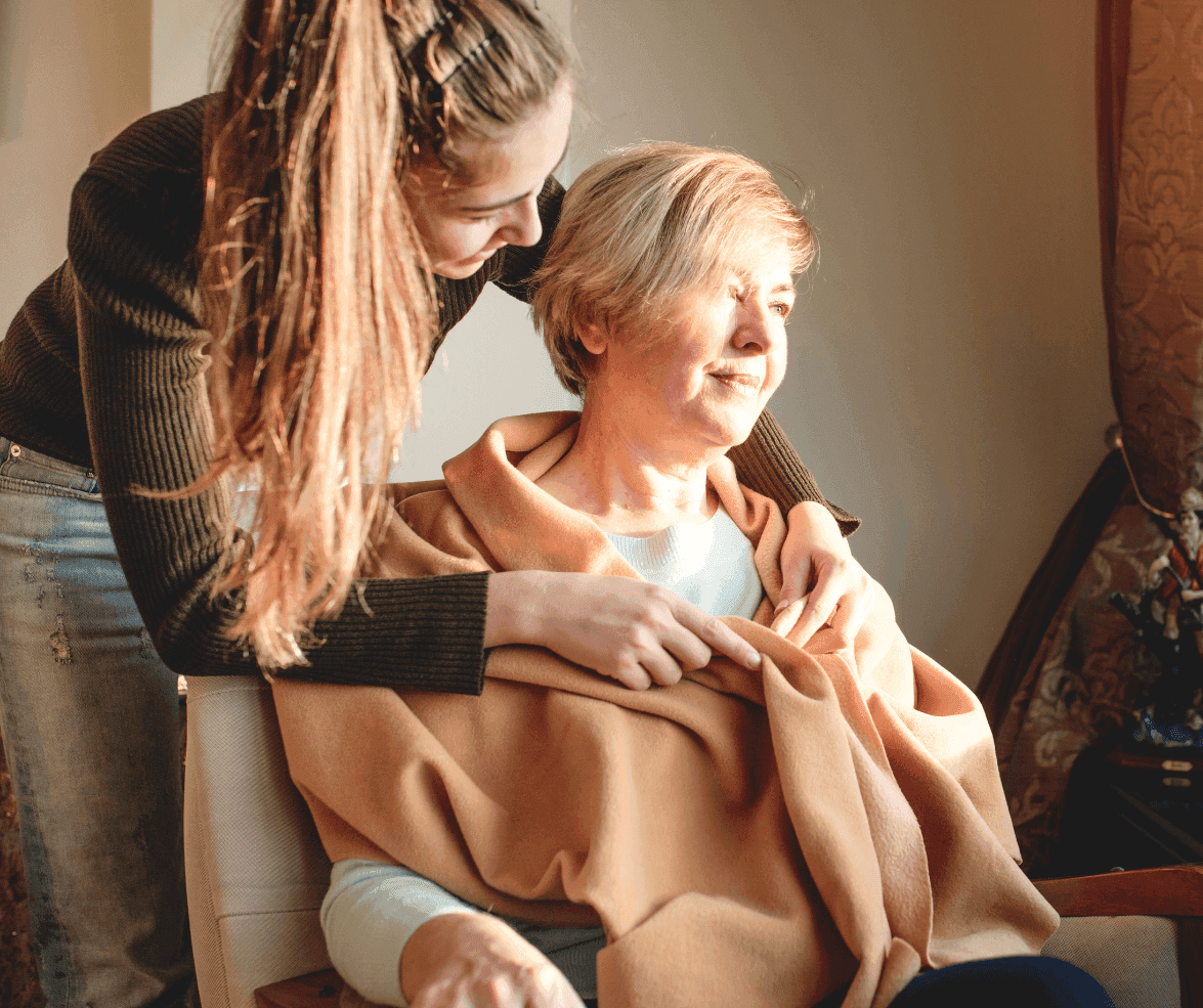 Young woman drapes a cozy blanket around an older woman who is sitting and smiling warmly. - Home Instead