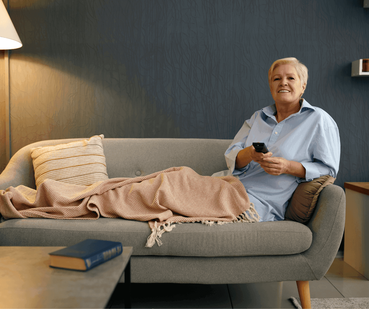 Person relaxing on a sofa with a blanket, holding a remote control, and smiling in a cozy living room. - Home Instead