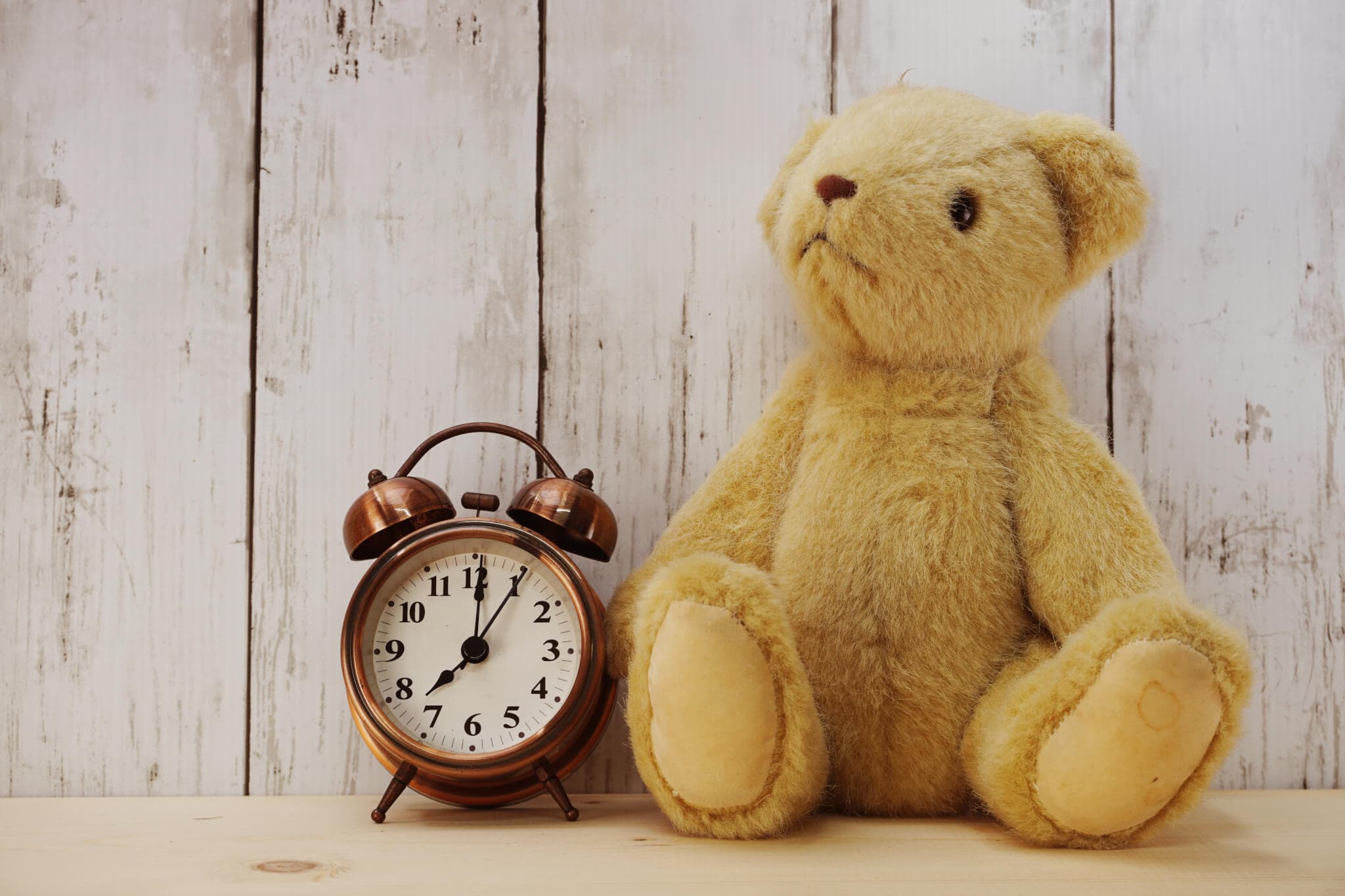 A teddy bear and a vintage alarm clock sit on a wooden surface against a rustic wooden wall. - Home Instead