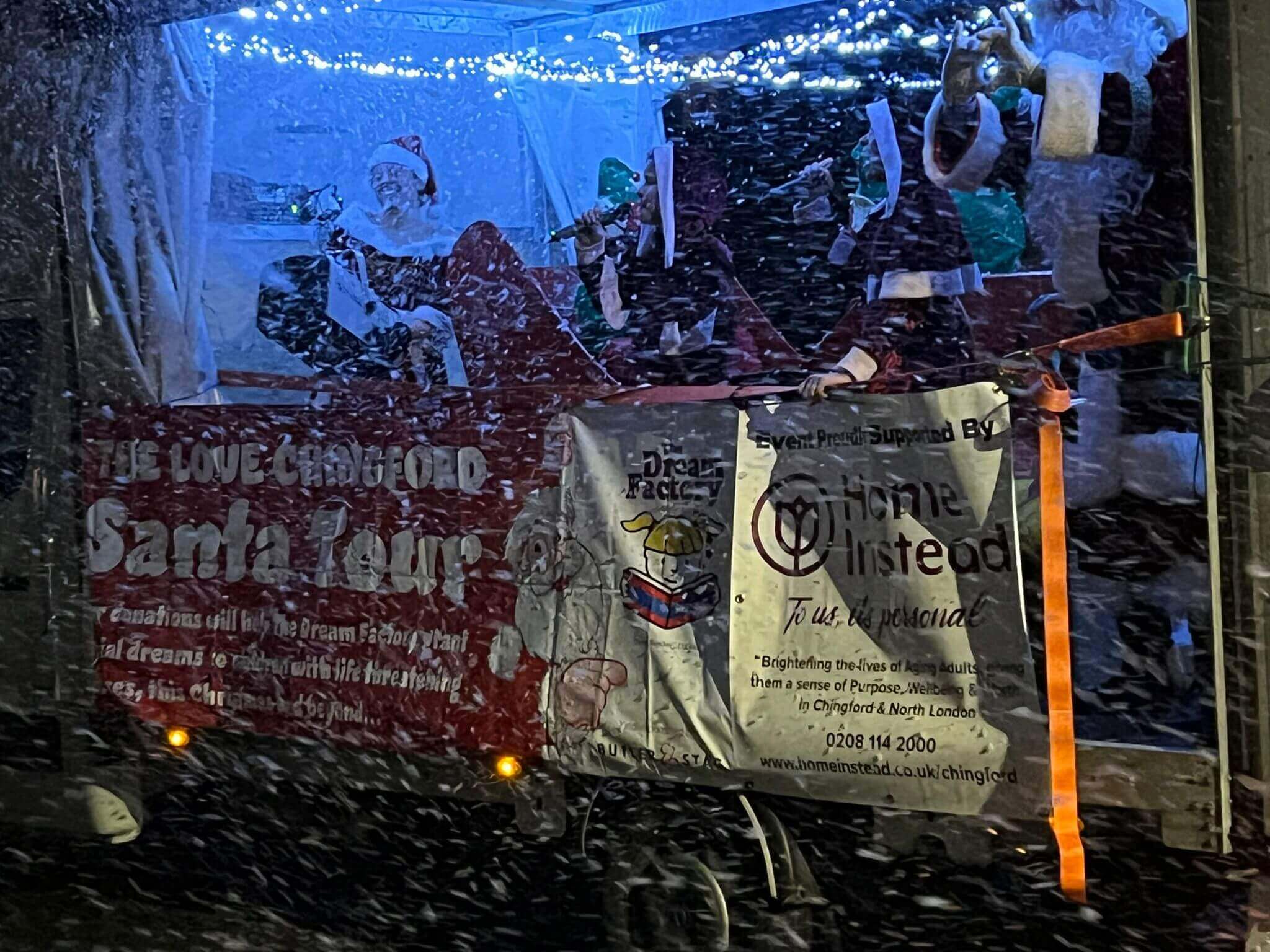 A festive float in snowfall with Santa Claus, lights, and banners reading "Santa Tour" and "Chimneylins". - Home Instead