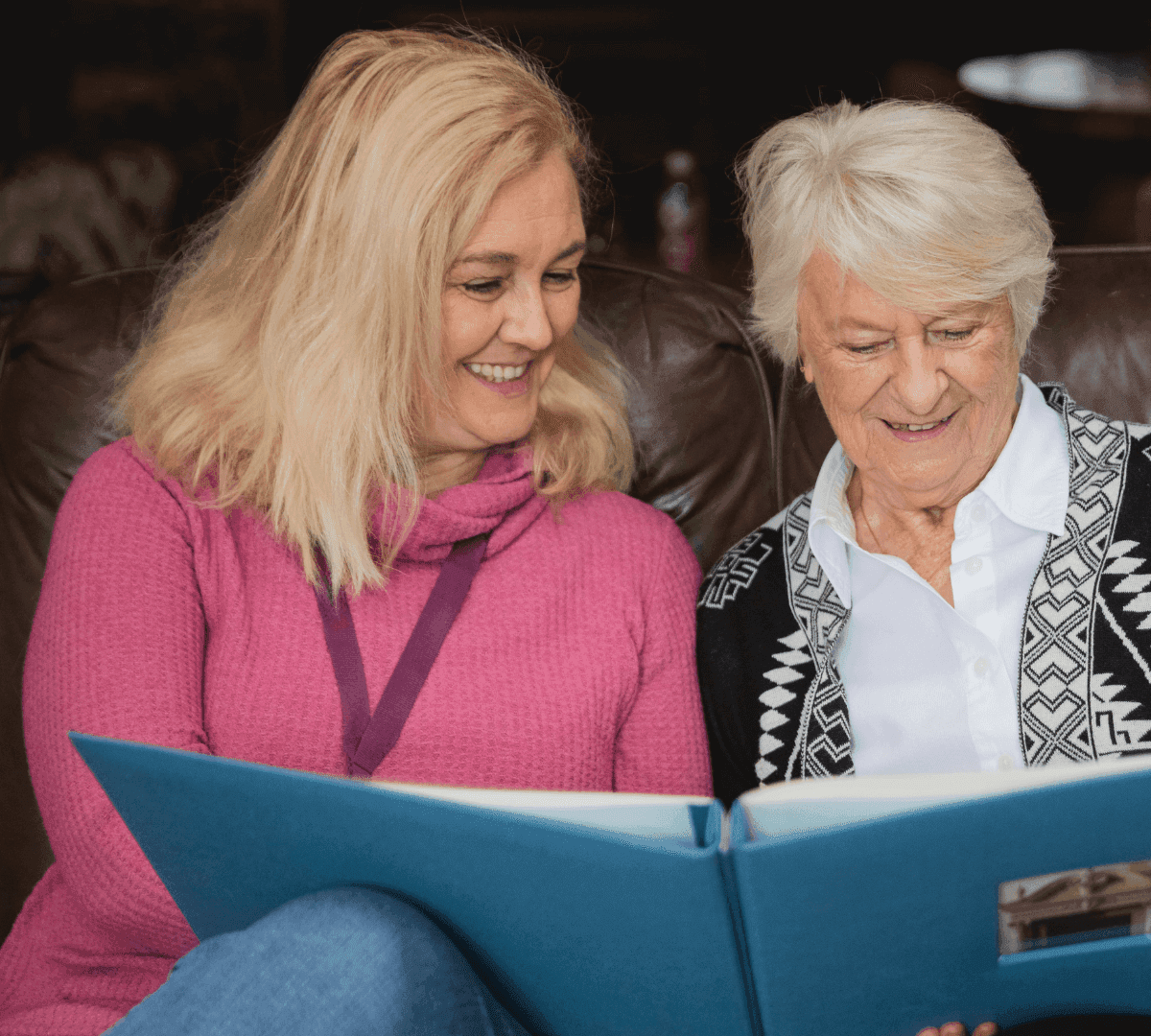 Two women, one elderly and one younger, smiling while looking at a large blue photo album together on a couch. - Home Instead