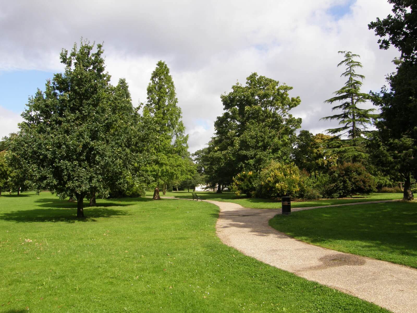 A paved pathway winds through a lush park with green grass, trees, and a partly cloudy sky. - Home Instead