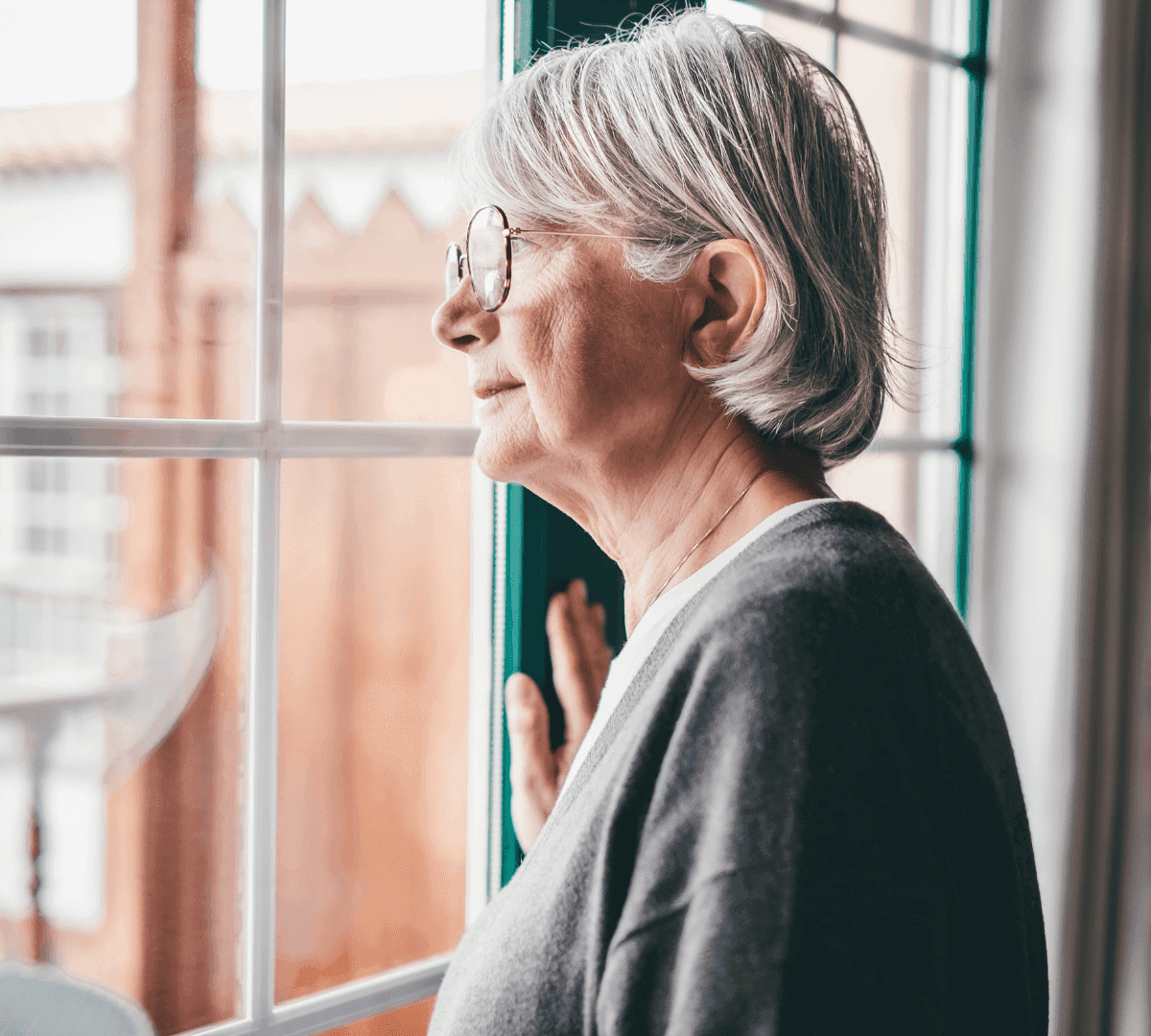 Elderly woman with glasses and gray hair looking out of a window with a serene expression. - Home Instead