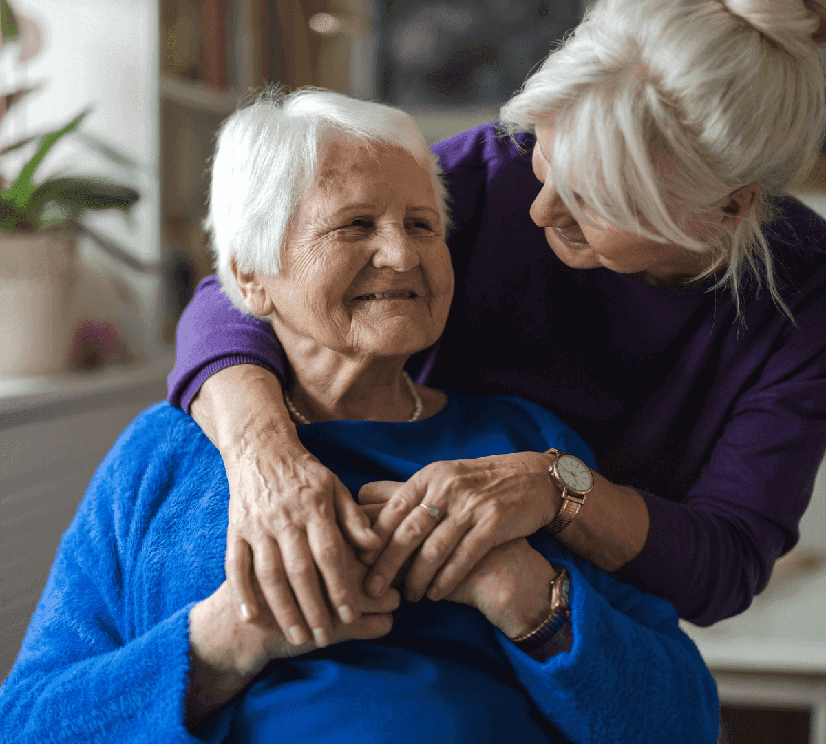 Two elderly women share a warm embrace, one sitting and smiling while the other stands, arms draped affectionately around her. - Home Instead