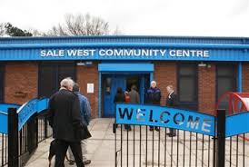 People entering a blue and red brick building labeled "Sale West Community Centre," with a "Welcome" sign on the gate. - Home Instead