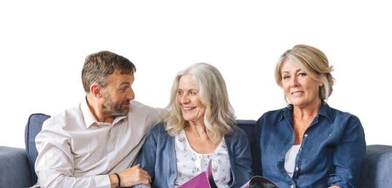 Three people sitting on a couch, one man on the left and two women, smiling and looking at each other, holding a book. - Home Instead