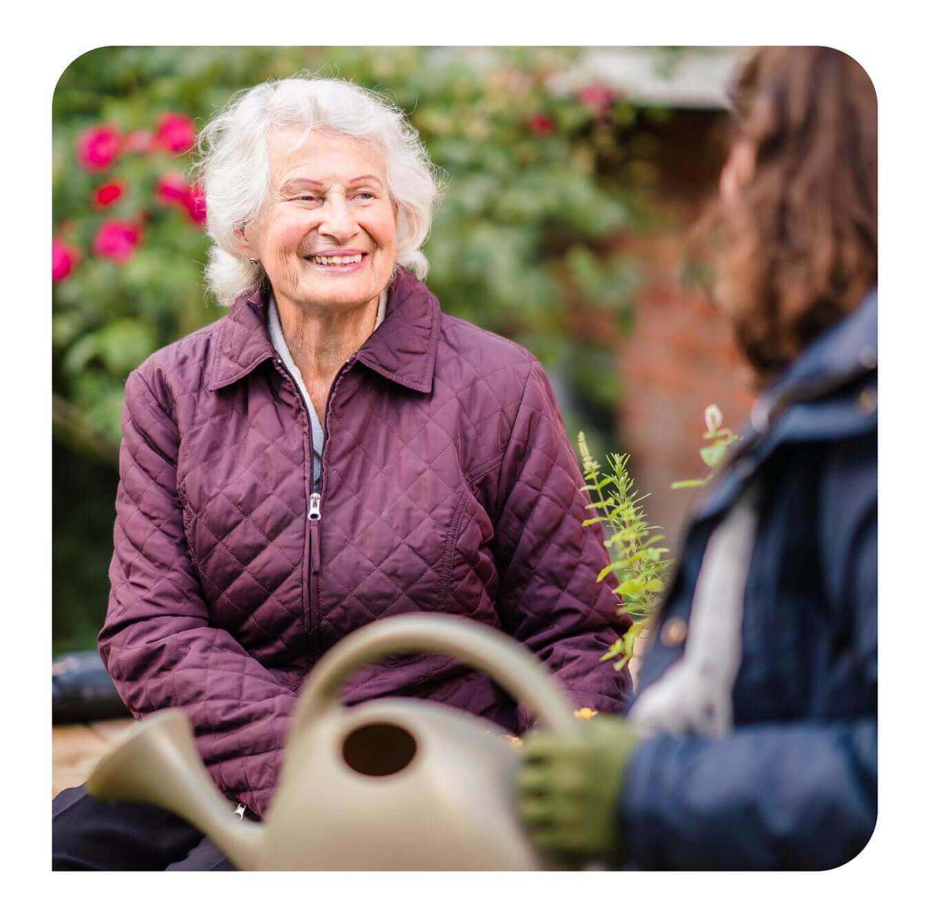 An elderly woman smiles while sitting in a garden, engaging in conversation with another person holding a watering can. - Home Instead