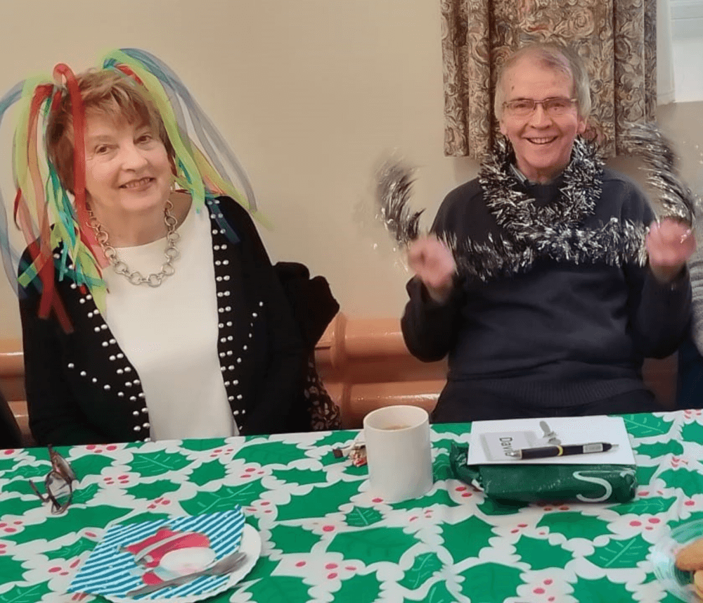Two smiling people wearing festive accessories sit at a table with holiday decorations and a cup of coffee. - Home Instead